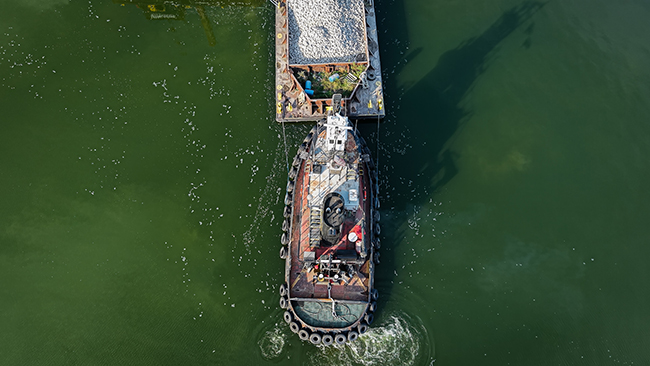 An aerial photo taken directly above a tug surrounded by water. The tug is egg shaped and has a row of tires lining the entire perimeter. An American flag is visible on the tug. Ahead of the tug is the back end of a barge loaded with cobble. The motors of the tug are churning the water.
